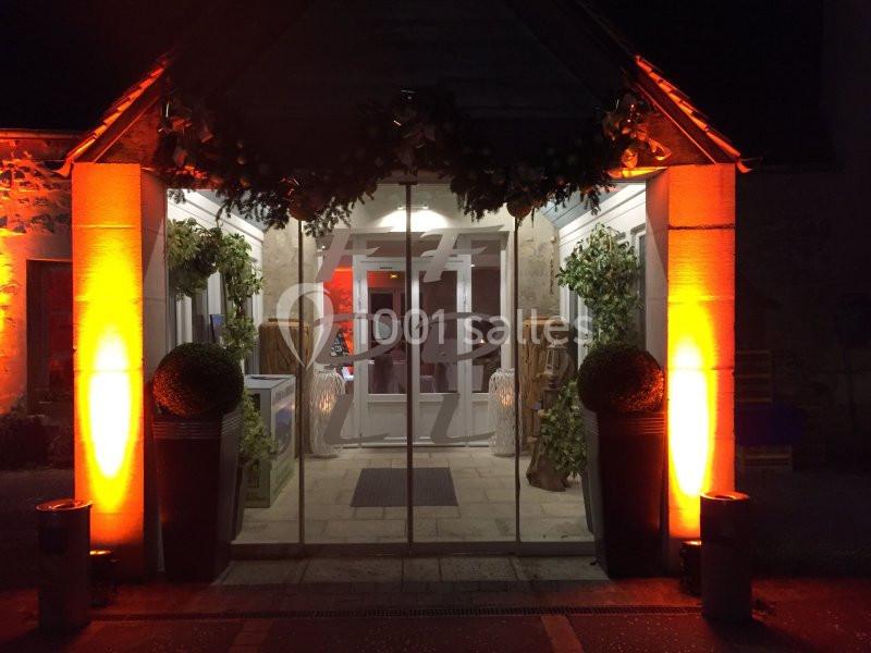 Entrée vitrée illuminée par des projecteurs orange, décorée de plantes et de guirlandes florales.