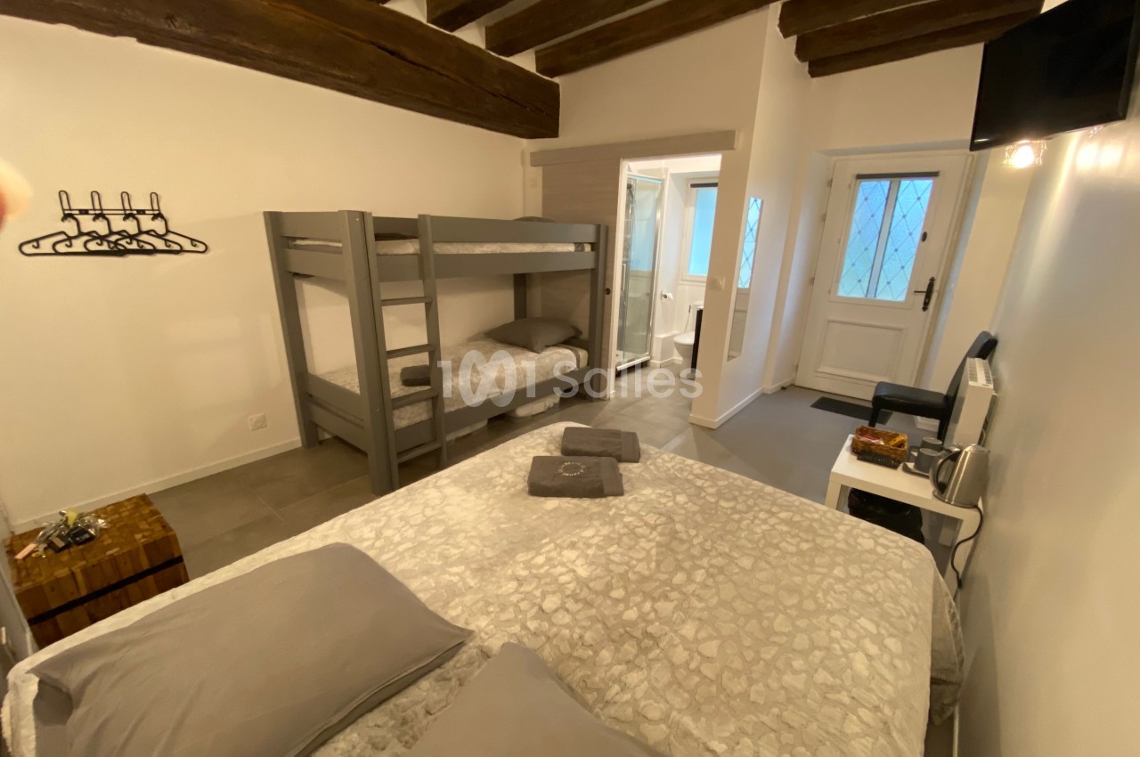 Chambre avec lit double, lits superposés, poutres apparentes, salle de bain attenante et porte-fenêtre.
