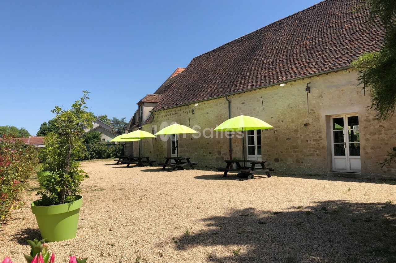 Cour extérieure avec tables en bois et parasols verts, devant un bâtiment en pierre sous un ciel dégagé.