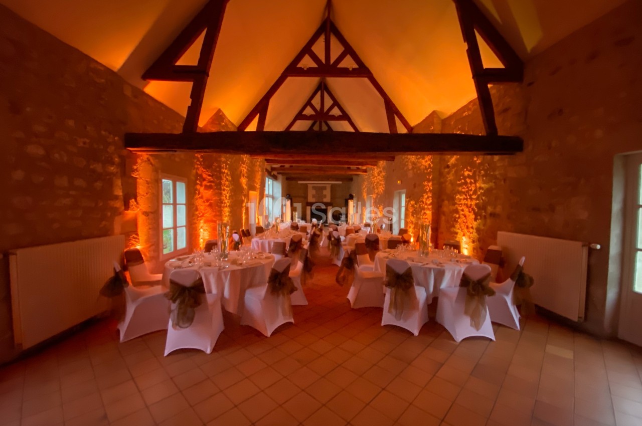Salle de réception avec tables rondes dressées, éclairage chaleureux et poutres apparentes en bois.