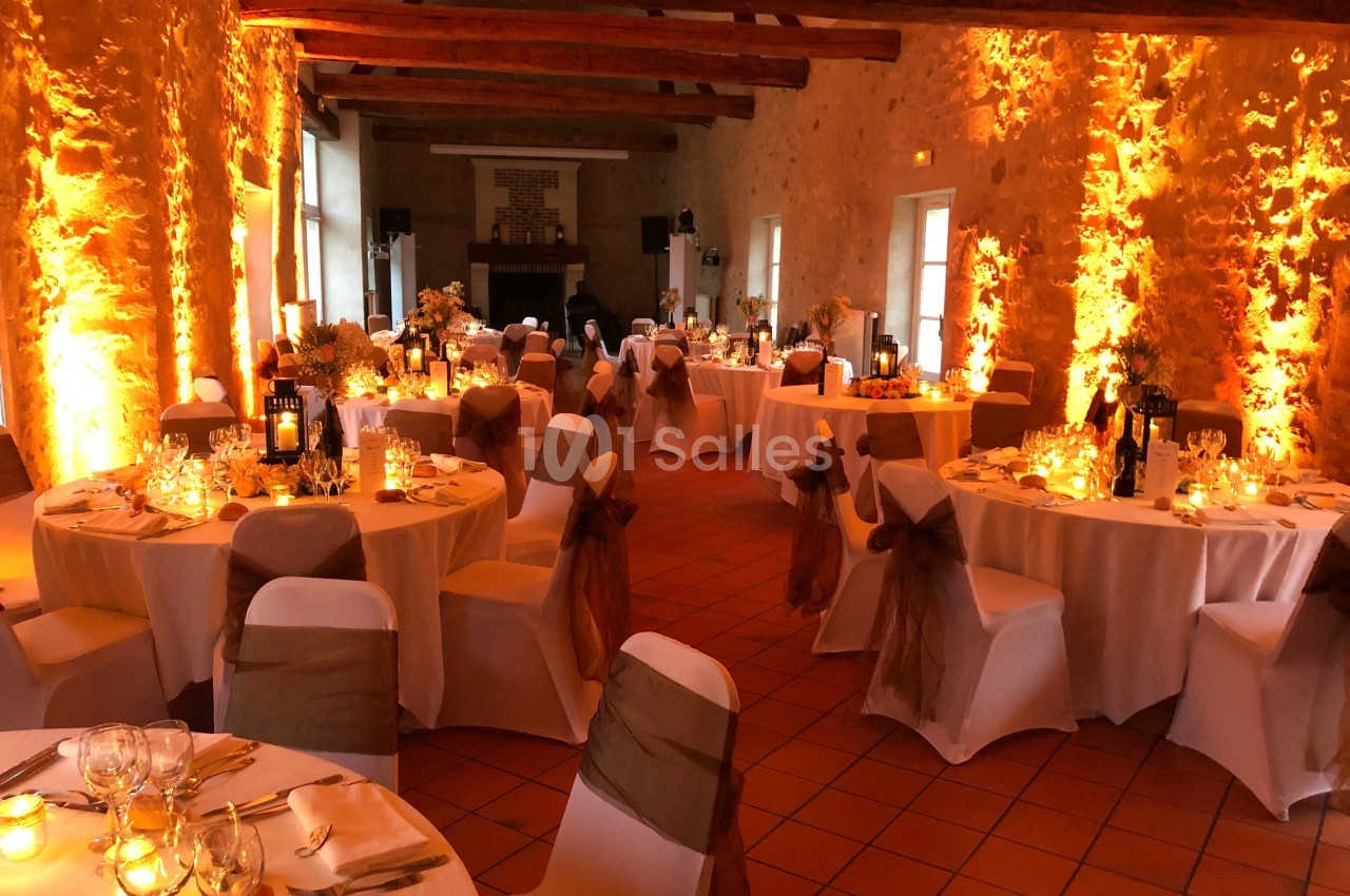 Salle de réception décorée avec des tables rondes, nappes blanches, chaises ornées de rubans et éclairage chaleureux.