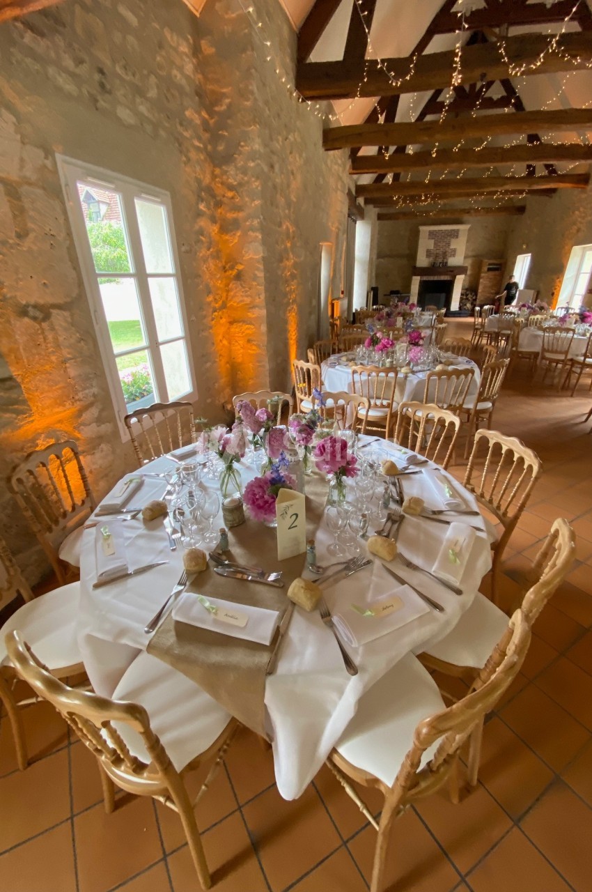 Salle de réception décorée avec des tables rondes dressées, ornées de fleurs et éclairées par des guirlandes lumineuses.