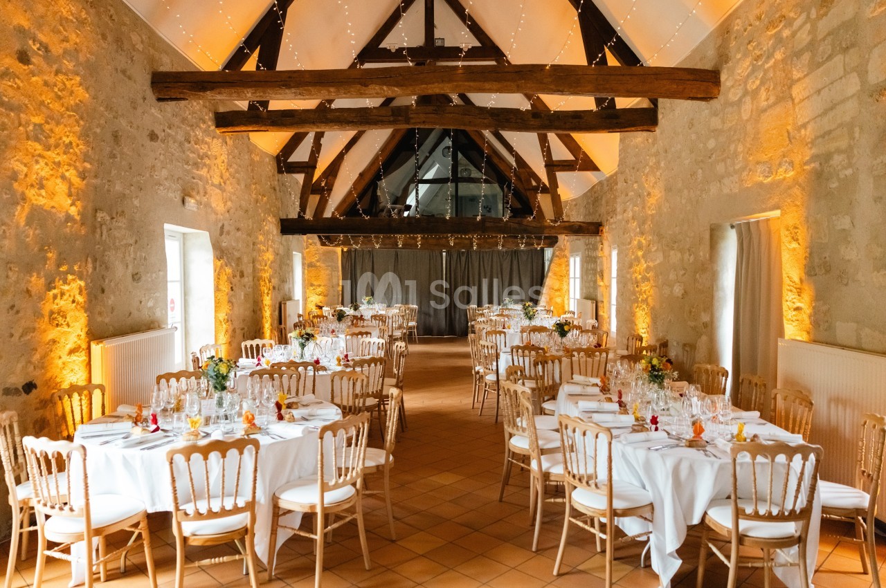 Salle de réception avec tables rondes dressées, chaises en bois clair et éclairage chaleureux sous une charpente apparente.