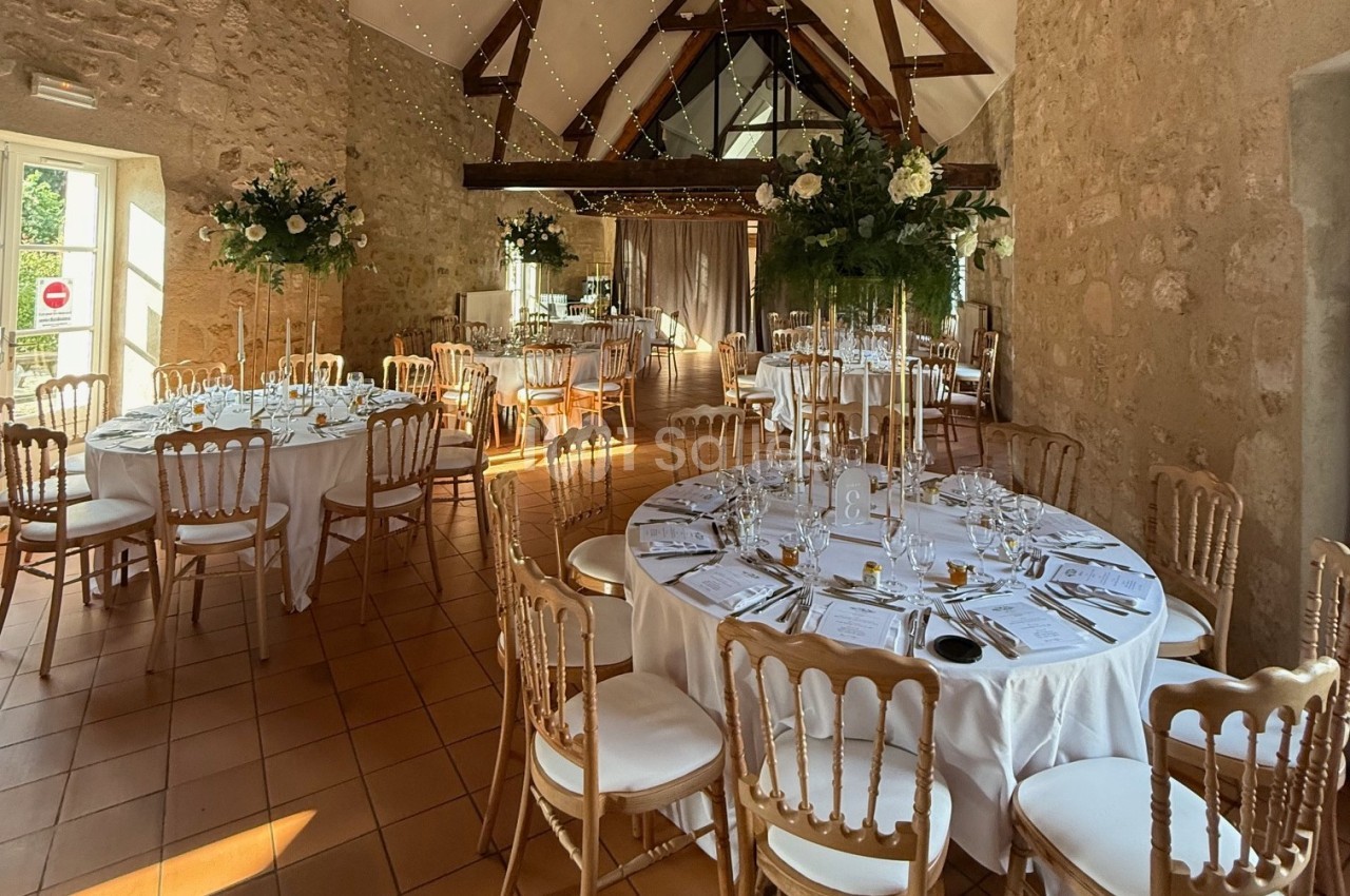 Salle de réception décorée pour un mariage avec tables rondes dressées, chaises dorées et arrangements floraux.