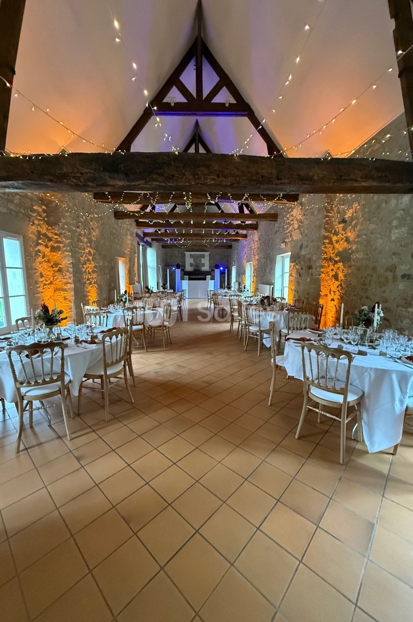 Salle de réception avec tables dressées, chaises en bois clair, murs en pierre et guirlandes lumineuses suspendues.