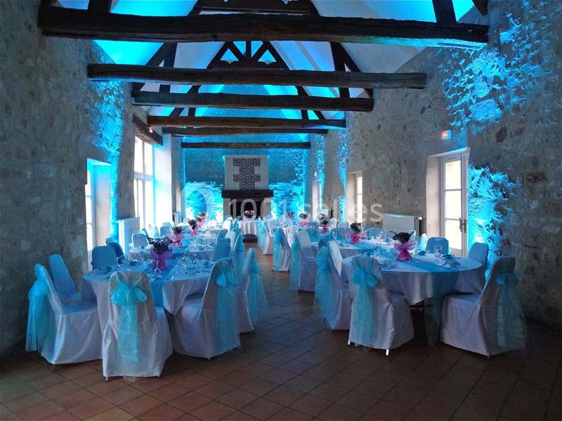 Salle de réception en pierre avec tables rondes décorées de nappes blanches et nœuds bleus, éclairage bleu tamisé.