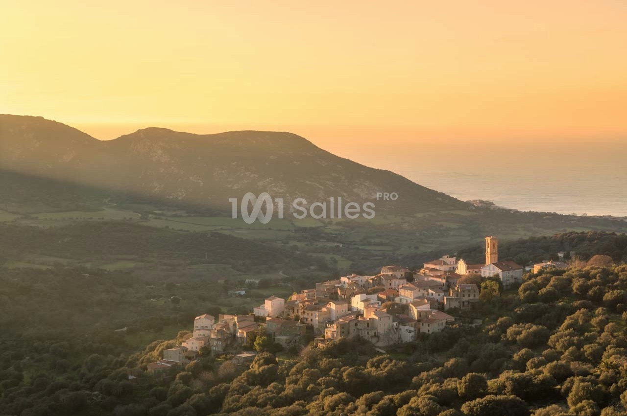 Village perché entouré de végétation, baigné par une lumière dorée au coucher du soleil, avec des collines en arrière-plan.