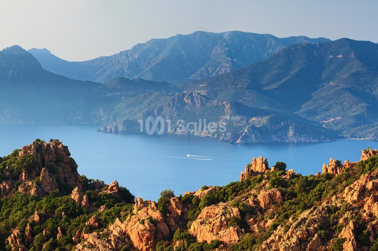 Paysage côtier avec des formations rocheuses rouges au premier plan, une mer bleue et des montagnes verdoyantes à l'arrière…