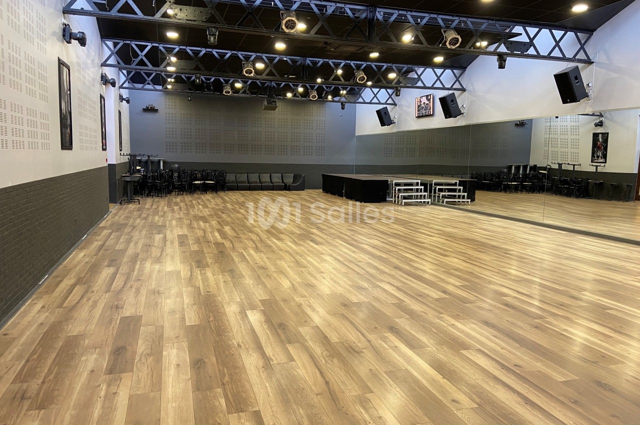 Salle spacieuse avec parquet en bois, murs clairs, miroirs et éclairage suspendu, équipée de chaises et d'une estrade.