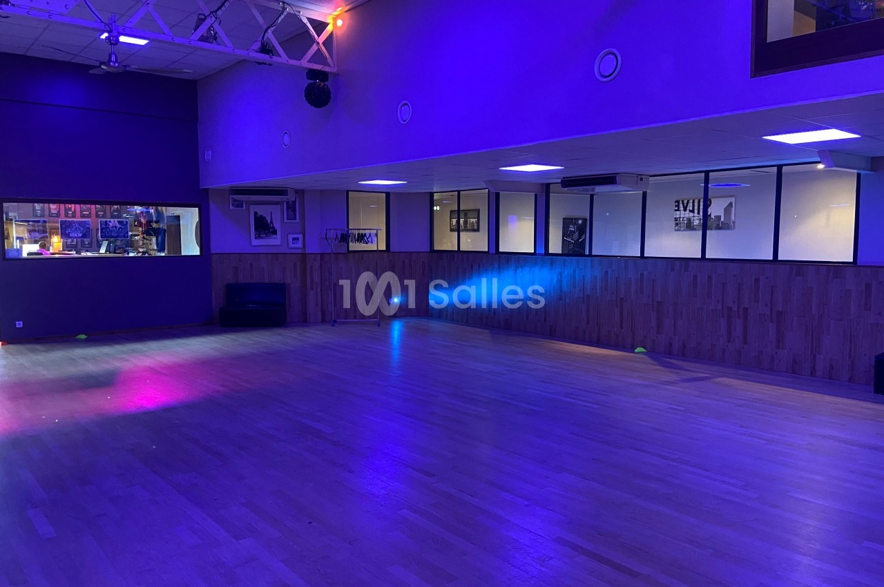 Salle de danse vide avec parquet clair, éclairage tamisé bleu et violet, et murs vitrés donnant sur un espace adjacent.