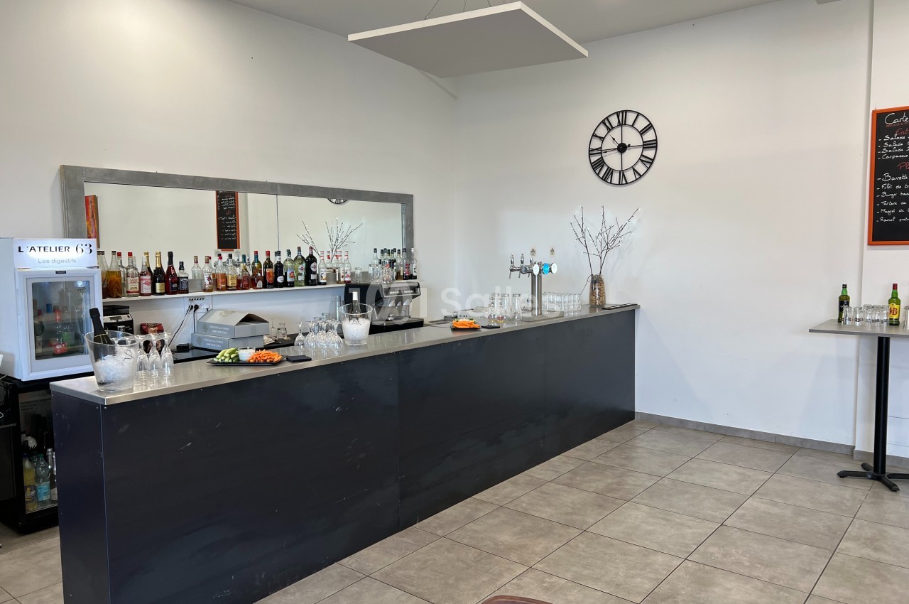 Comptoir de bar avec bouteilles alignées, verres, machine à café et décor sobre dans une pièce lumineuse.