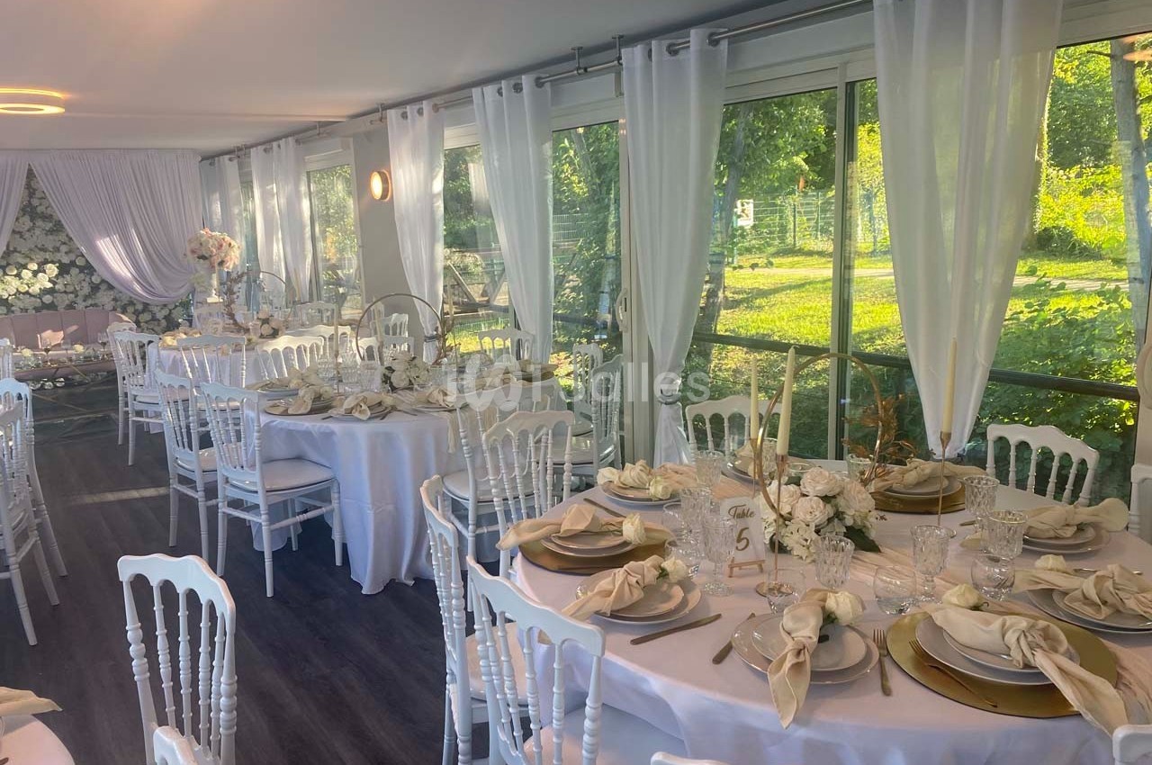 Salle de réception lumineuse avec tables décorées de nappes blanches, vaisselle élégante et chaises blanches, vue sur jardin.