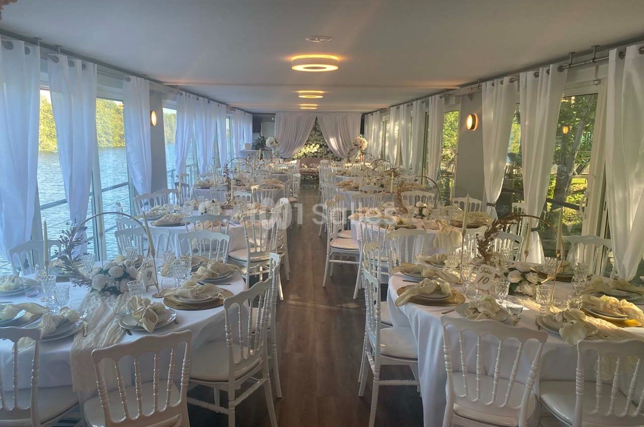 Salle de réception élégamment décorée avec des tables rondes, nappes blanches et chaises disposées près de grandes baies…