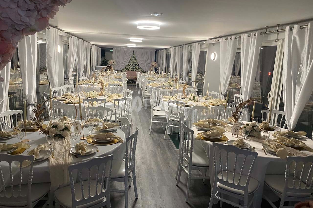 Salle de réception décorée avec des tables rondes dressées, nappes blanches, vaisselle élégante et rideaux translucides.