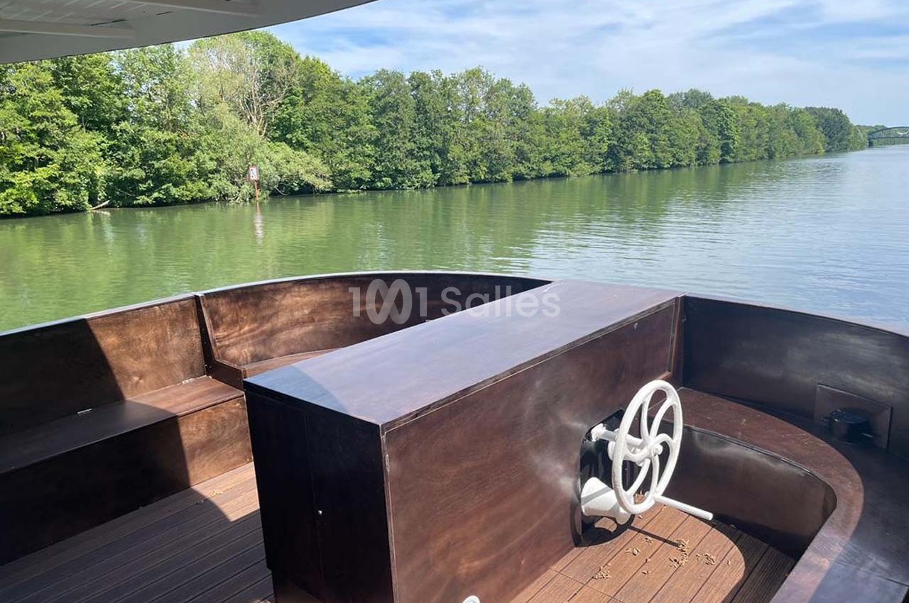 Vue d'un bateau en bois avec une barre de navigation, naviguant sur une rivière bordée d'arbres sous un ciel dégagé.