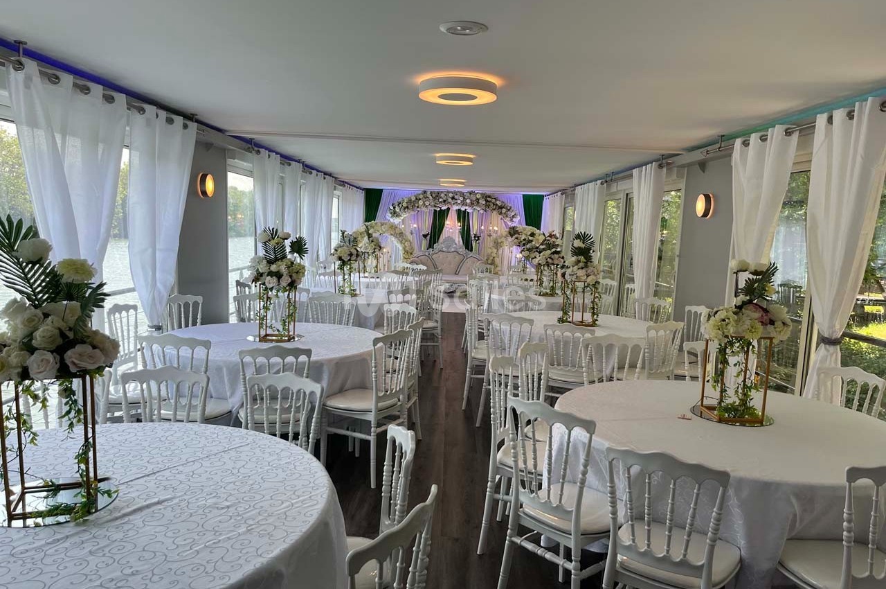 Salle de réception élégante avec tables rondes décorées de fleurs blanches et chaises blanches, éclairée par la lumière…