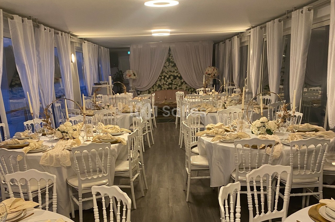 Salle de réception élégamment décorée avec des tables rondes, nappes blanches, chaises assorties et arrangements floraux.