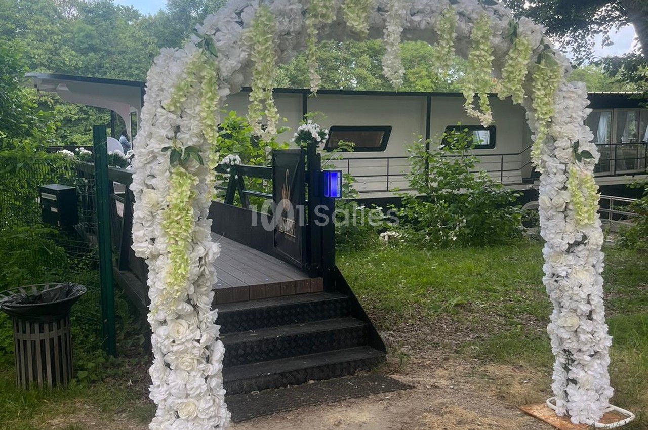 Arche décorée de fleurs blanches et vertes devant une passerelle menant à un bâtiment entouré de verdure.