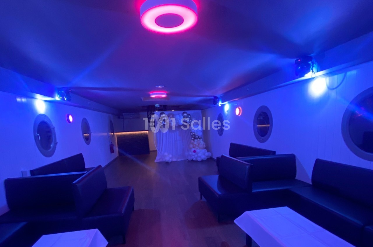 Salle intérieure d'un bateau avec éclairage tamisé bleu et rose, banquettes noires et décorations en ballons au fond.