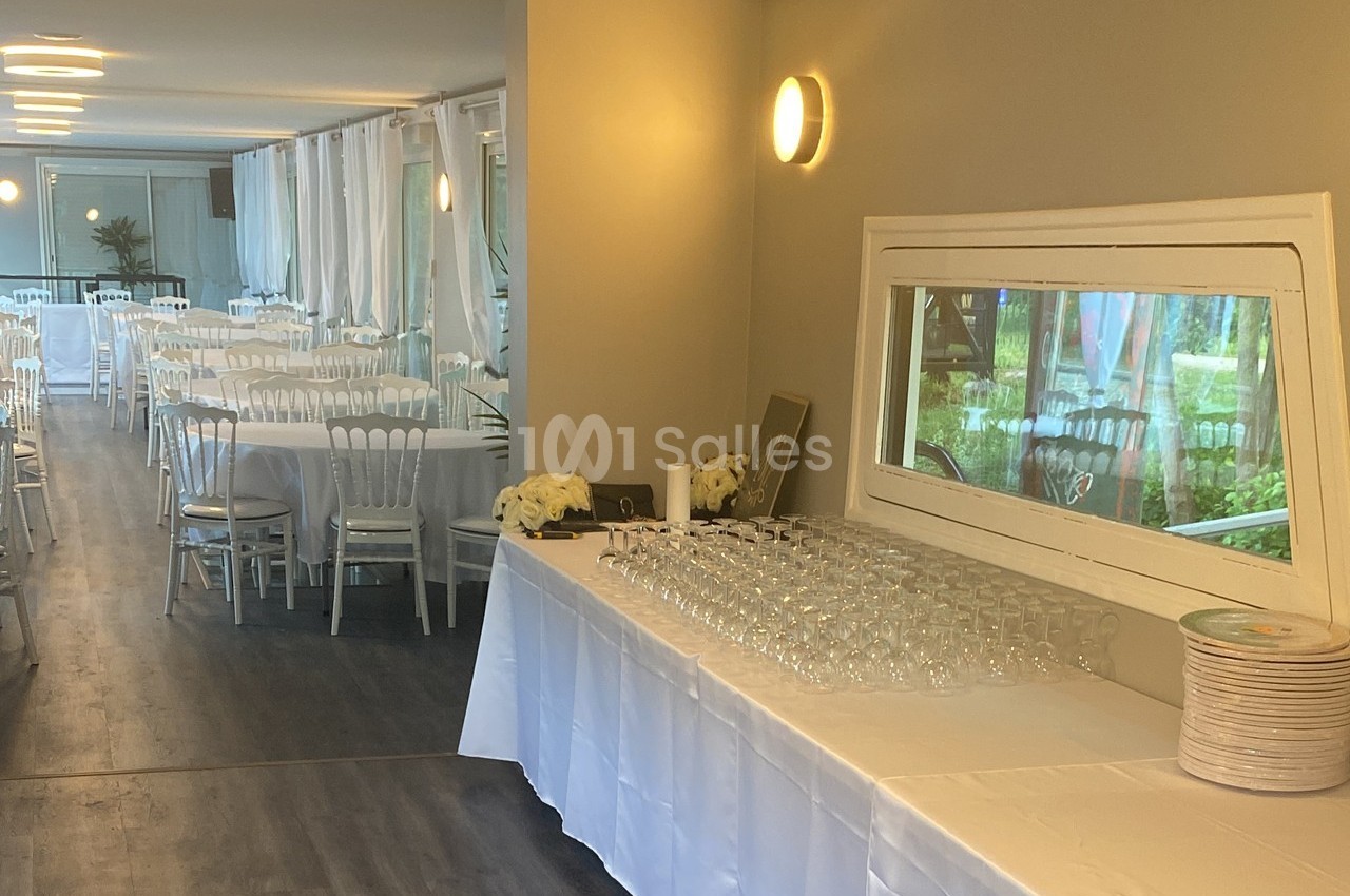 Salle de réception lumineuse avec tables dressées et un buffet de verres et assiettes sur une nappe blanche.