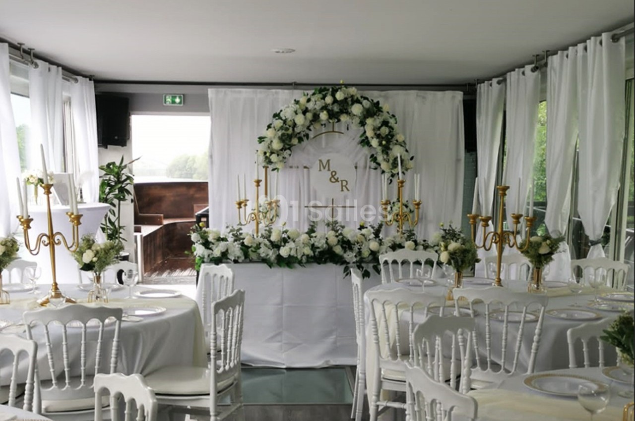Salle décorée pour un mariage avec des tables rondes, des chaises blanches, des chandeliers dorés et une arche florale.