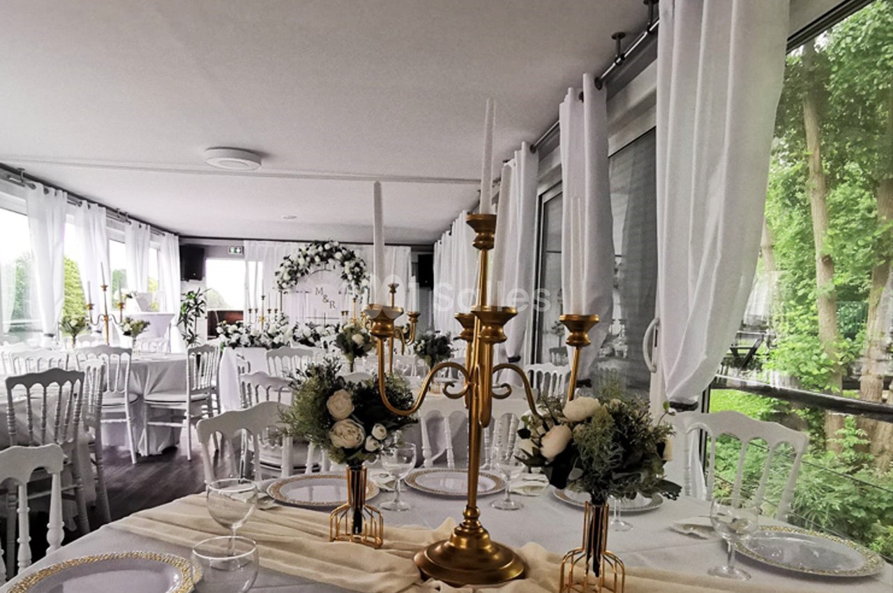 Salle de réception lumineuse avec tables décorées de chandeliers dorés, fleurs blanches et nappes élégantes.