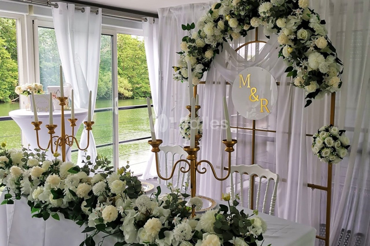 Table décorée pour un mariage avec chandeliers dorés, fleurs blanches et arche ornée des initiales ’M&R’, vue sur un lac.