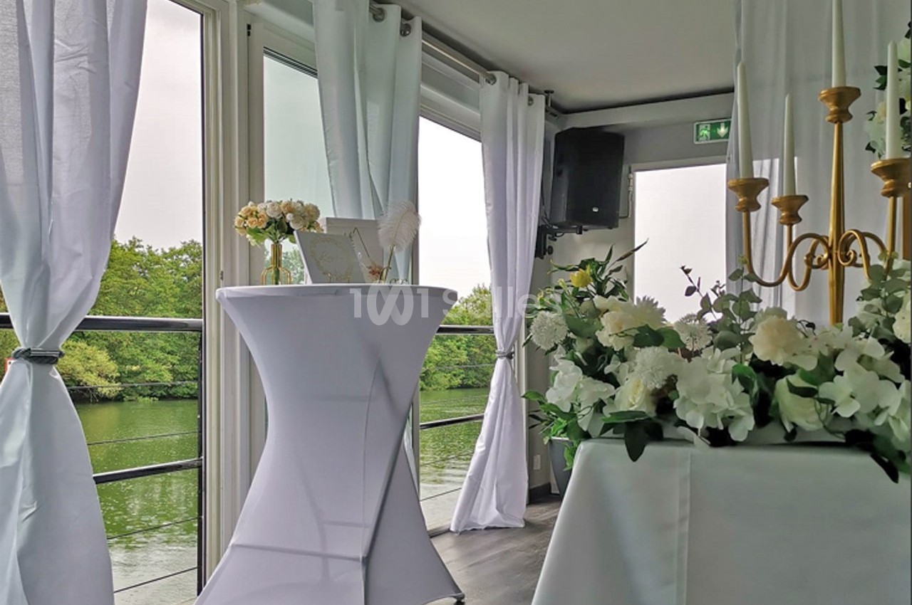 Salle lumineuse avec vue sur un plan d'eau, décorée de fleurs blanches, bougies et une table haute drapée de blanc.