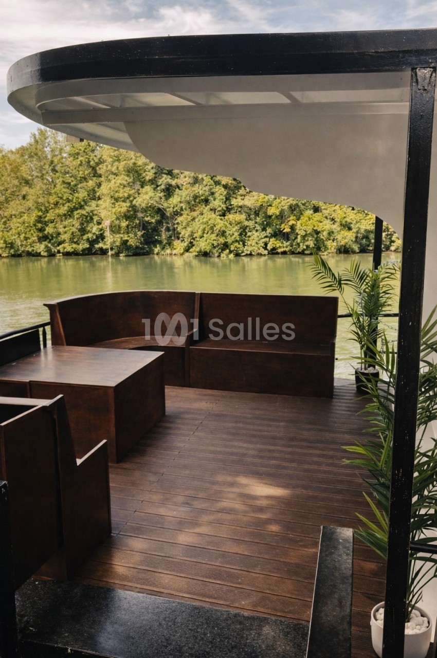 Terrasse en bois avec des bancs et des plantes, donnant sur une rivière bordée d'arbres sous un ciel partiellement nuageux.