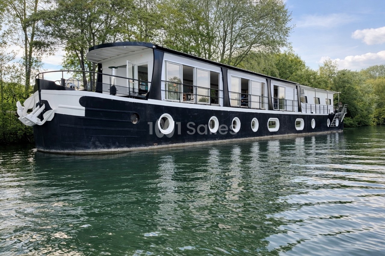 Péniche noire et blanche amarrée sur une rivière entourée d'arbres sous un ciel partiellement dégagé.