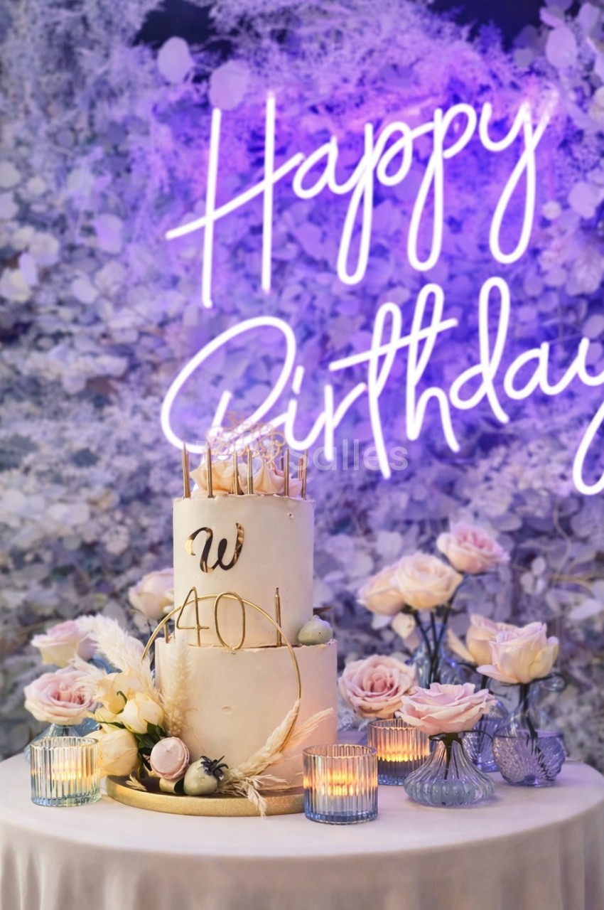 Gâteau d'anniversaire décoré de fleurs et bougies, entouré de roses, avec un néon ’Happy Birthday’ en arrière-plan.