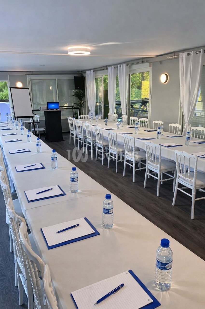Salle de réunion lumineuse avec tables alignées, chaises blanches, blocs-notes, bouteilles d'eau et écran de présentation.