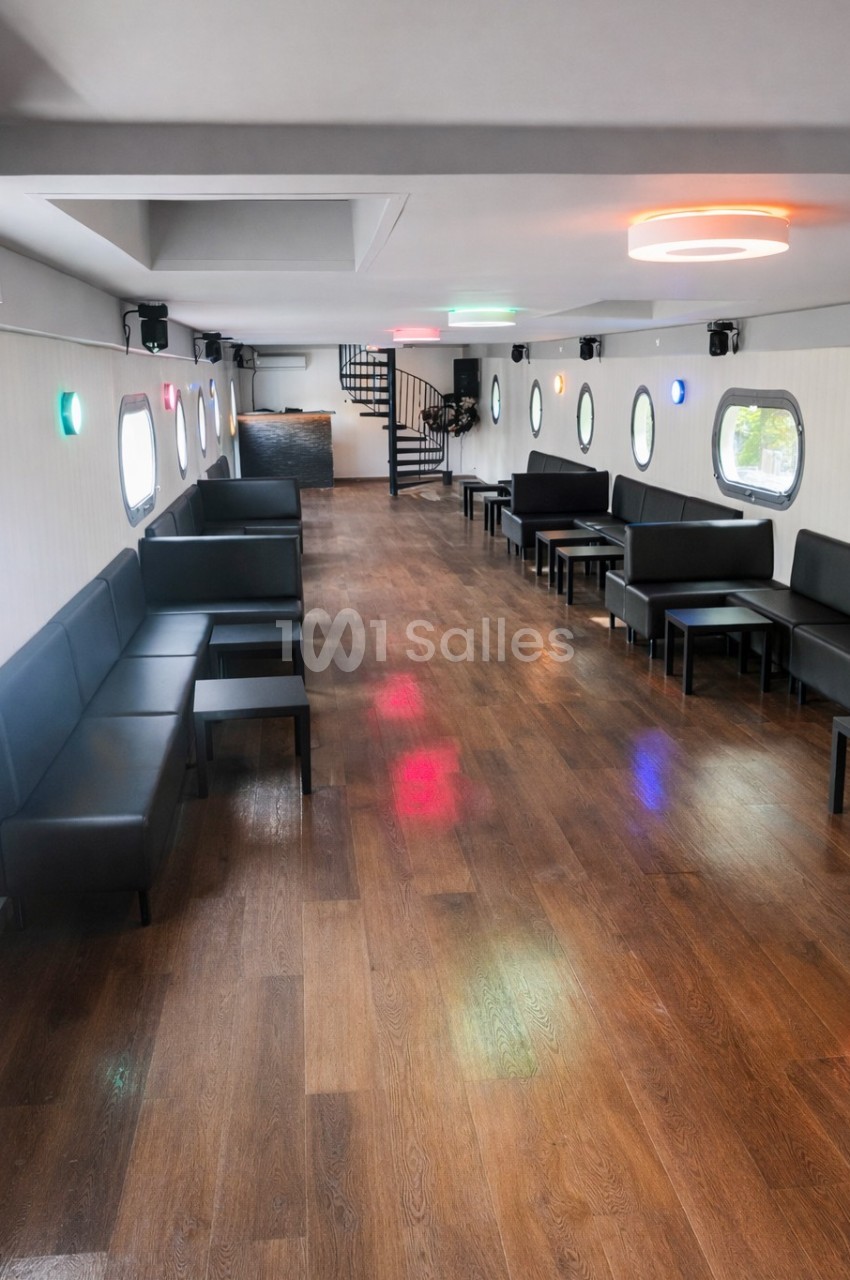 Salle intérieure lumineuse avec parquet en bois, banquettes noires, tables basses et hublots ronds sur les murs.