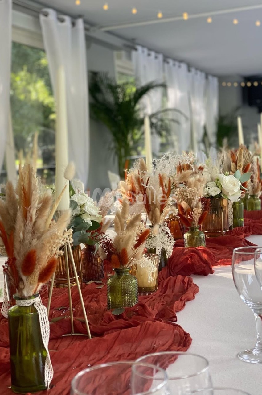 Table décorée avec des fleurs séchées, des bougies et des vases colorés sur un chemin de table rouge, dans une salle…
