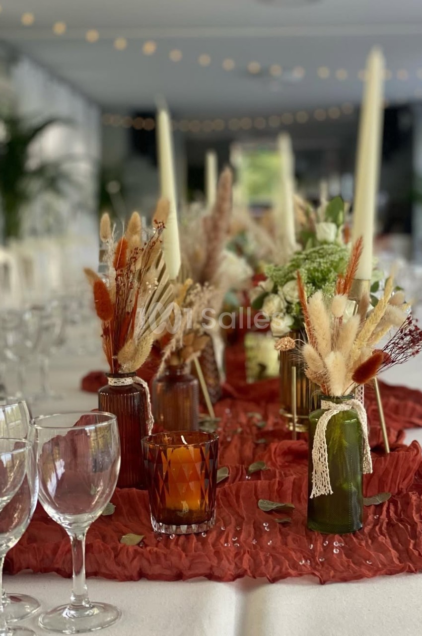 Décoration de table avec bougies, fleurs séchées et vases colorés sur une nappe rouge texturée.