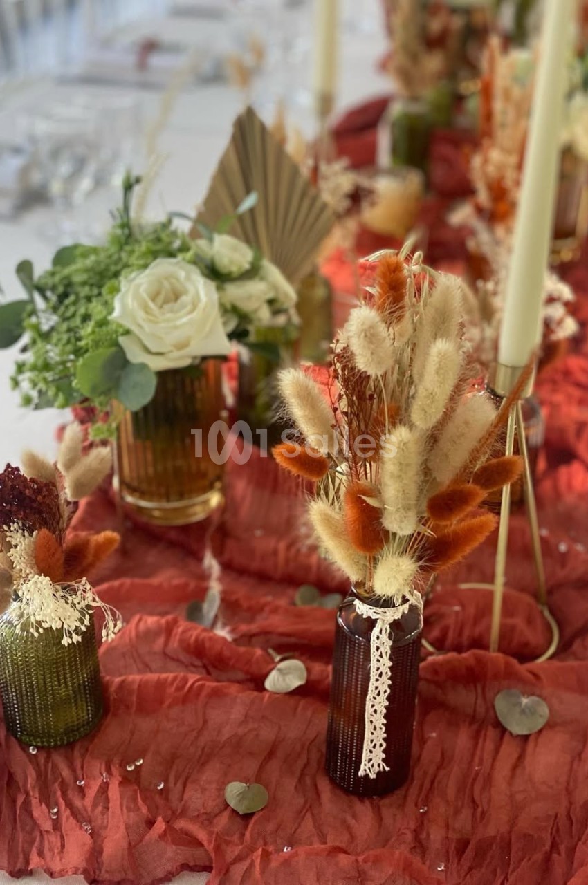 Décoration de table avec vases en verre, fleurs séchées et bougies, sur un tissu rouge plissé.