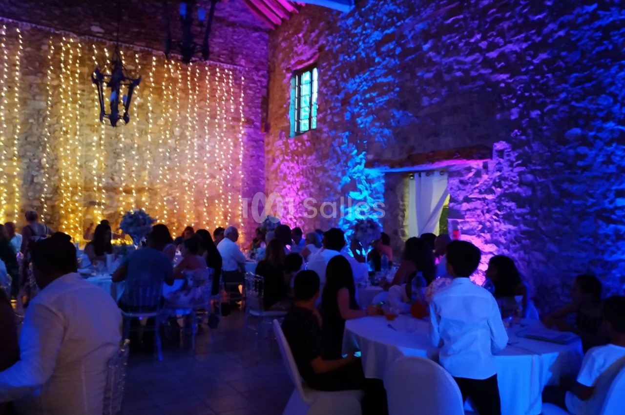 Salle en pierre éclairée par des lumières colorées, avec des invités assis à des tables lors d'un événement festif.