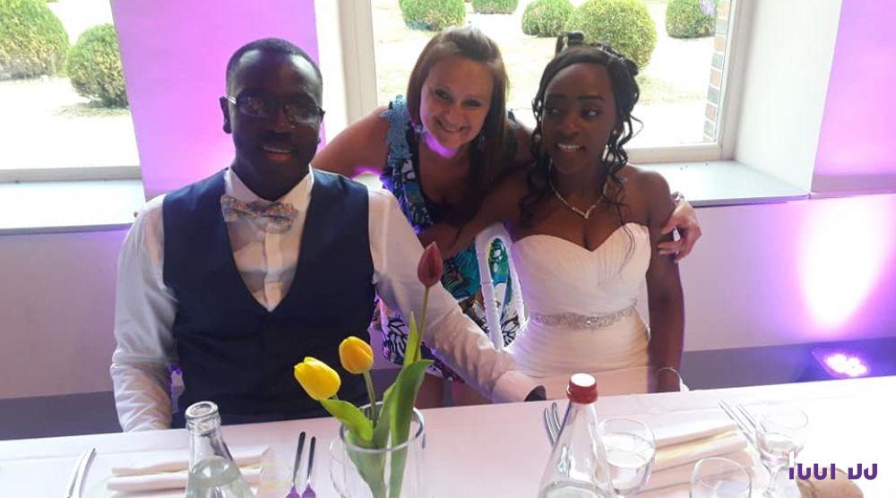 Un couple assis à une table de réception avec une femme debout derrière eux, des fleurs et des bouteilles au premier plan.