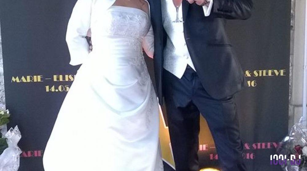 Un couple en tenue de mariage pose devant un panneau décoratif indiquant des prénoms et une date.