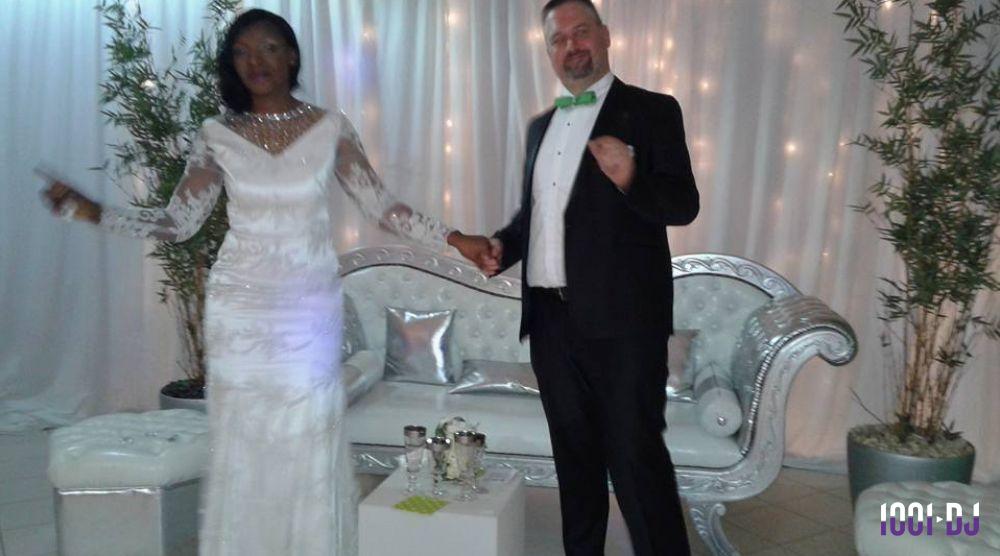 Un couple en tenue de mariage pose devant un canapé argenté, avec un décor lumineux et des plantes en arrière-plan.