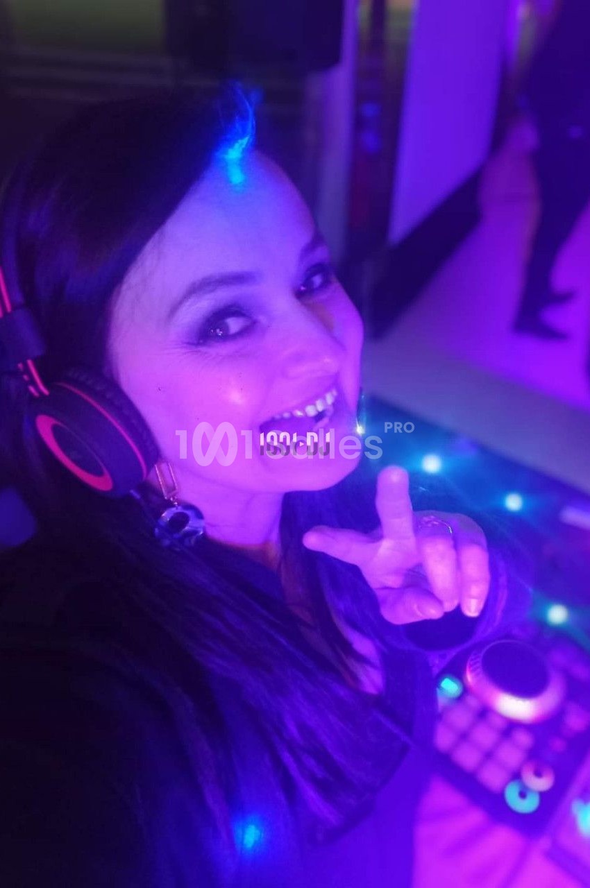 Femme portant un casque audio, souriante, faisant un signe de paix dans un environnement éclairé par des lumières colorées.