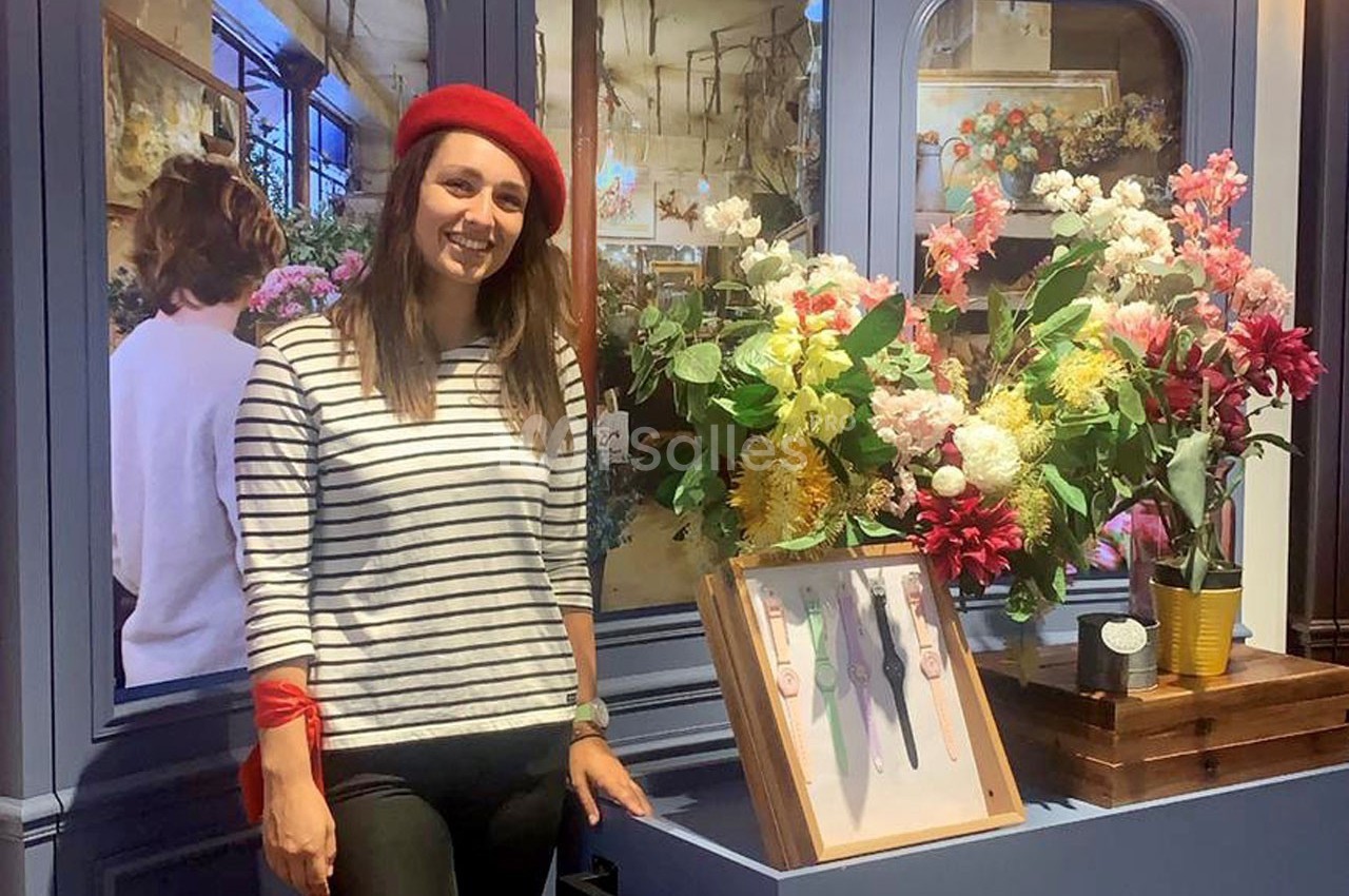 Femme souriante portant un béret rouge, debout près d'une vitrine décorée de fleurs colorées et de montres exposées.