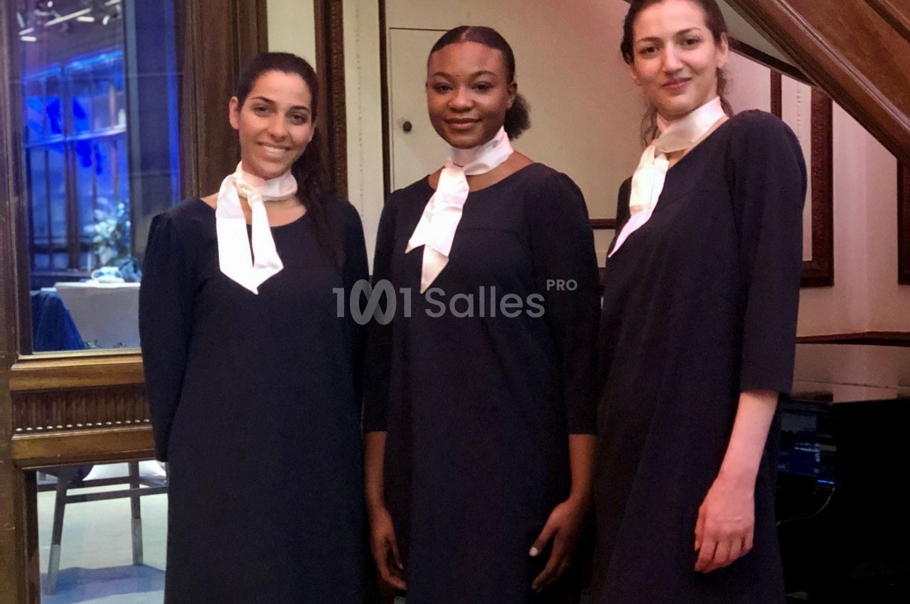 Trois femmes en tenue noire avec foulard blanc posent debout dans un intérieur élégant avec des cadres en bois.