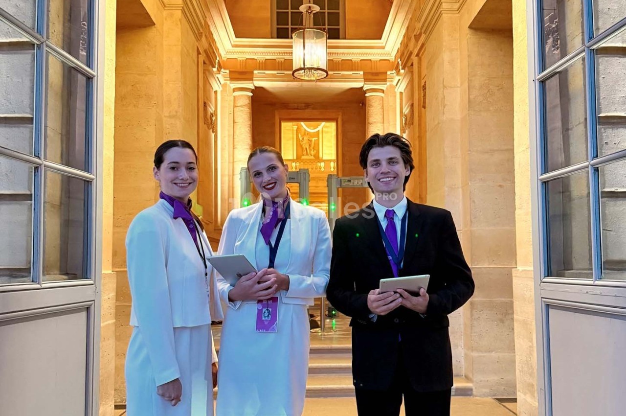 Trois hôtes souriants en tenue professionnelle, tenant des tablettes, devant une entrée lumineuse et élégante.
