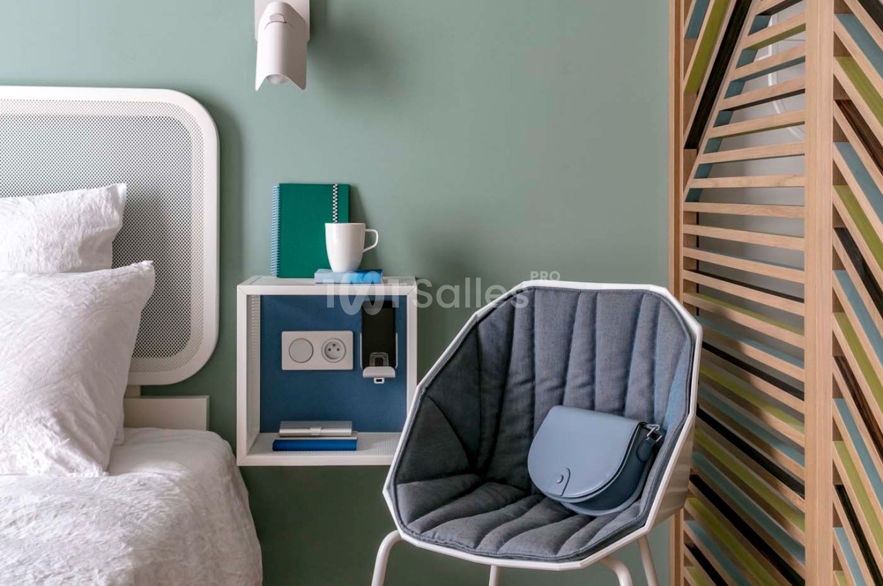 Coin de chambre avec lit, table de chevet intégrée, chaise grise avec sac bleu et cloison en bois colorée.