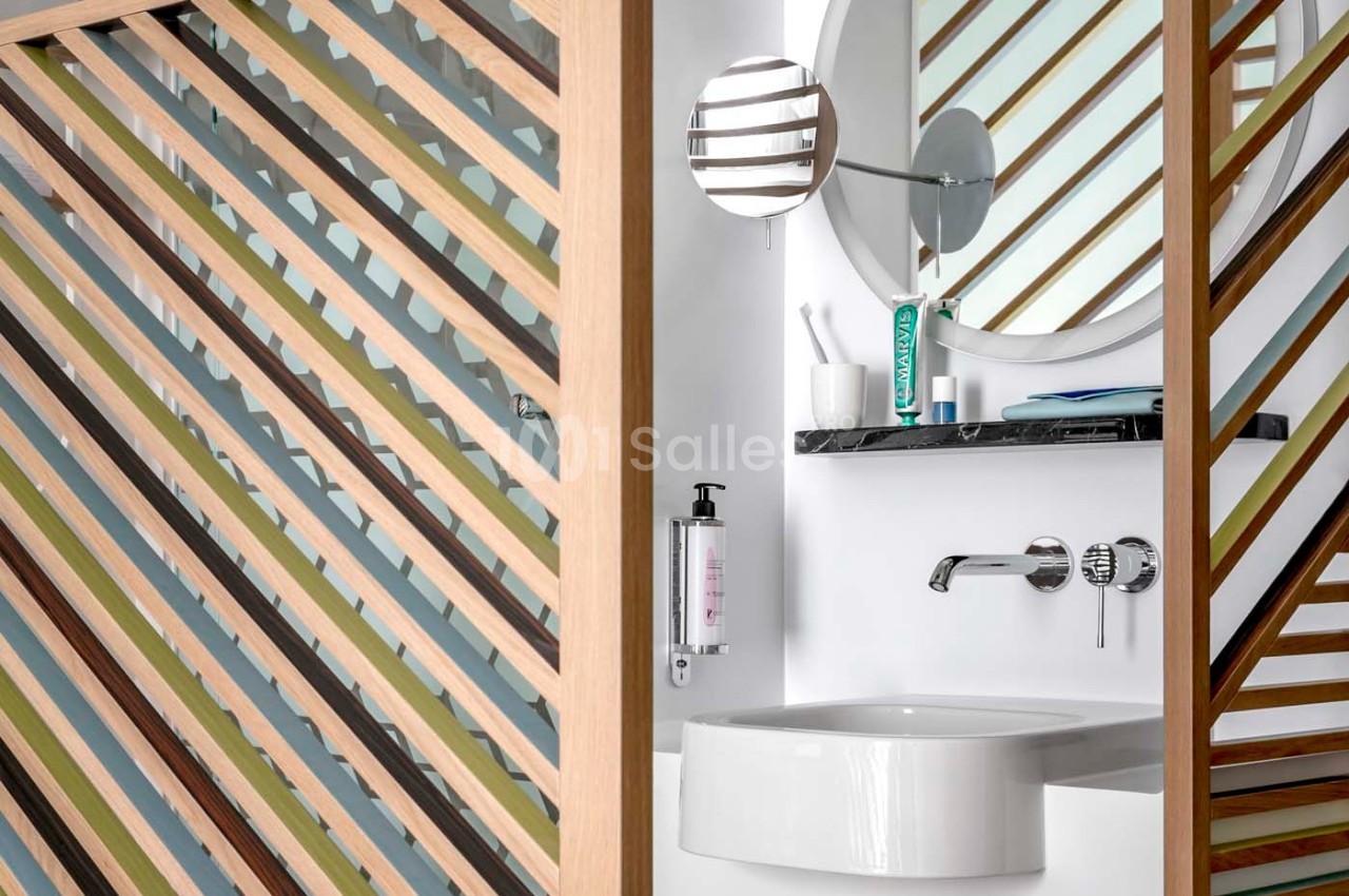 Lavabo moderne avec miroir rond, étagère murale, robinet chromé et panneaux en bois ajourés en premier plan.