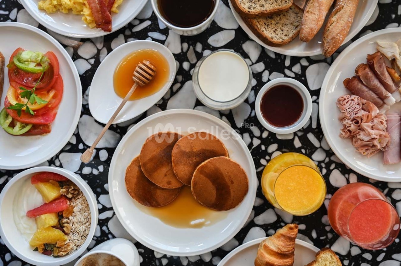 Petit-déjeuner varié avec pancakes, fruits, viennoiseries, charcuterie, pain, boissons et yaourt sur une table marbrée.