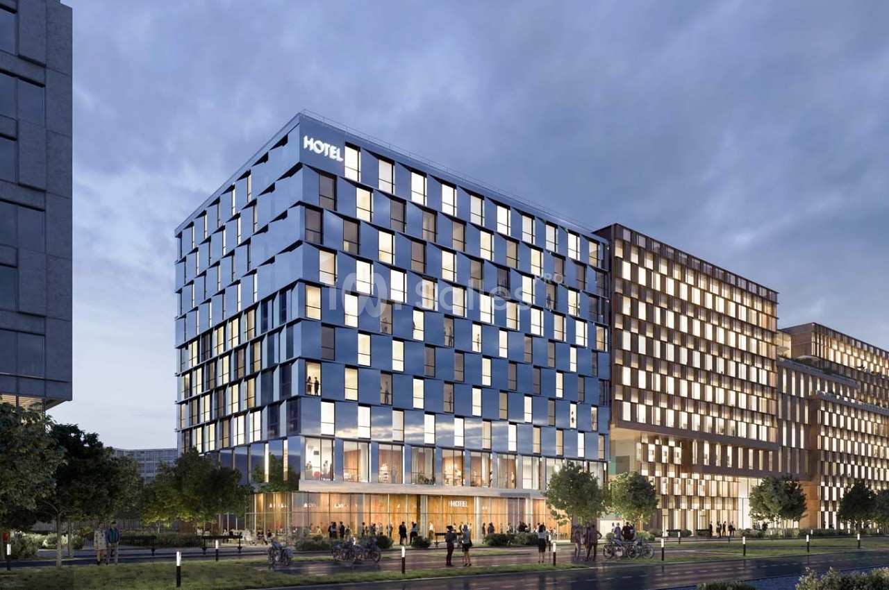 Façade moderne d'un hôtel en verre et métal, entourée d'immeubles et d'arbres, au crépuscule.