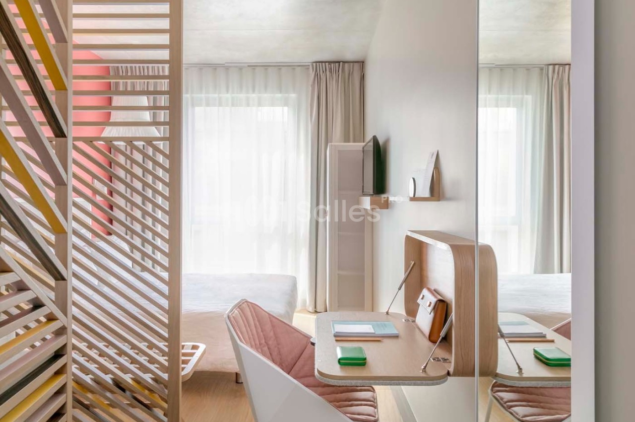 Bureau compact en bois clair avec chaise, étagères murales et lit en arrière-plan dans une pièce lumineuse.