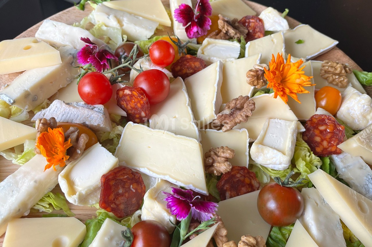 Plateau de fromages variés avec noix, tomates cerises, tranches de chorizo et fleurs comestibles sur un lit de salade.